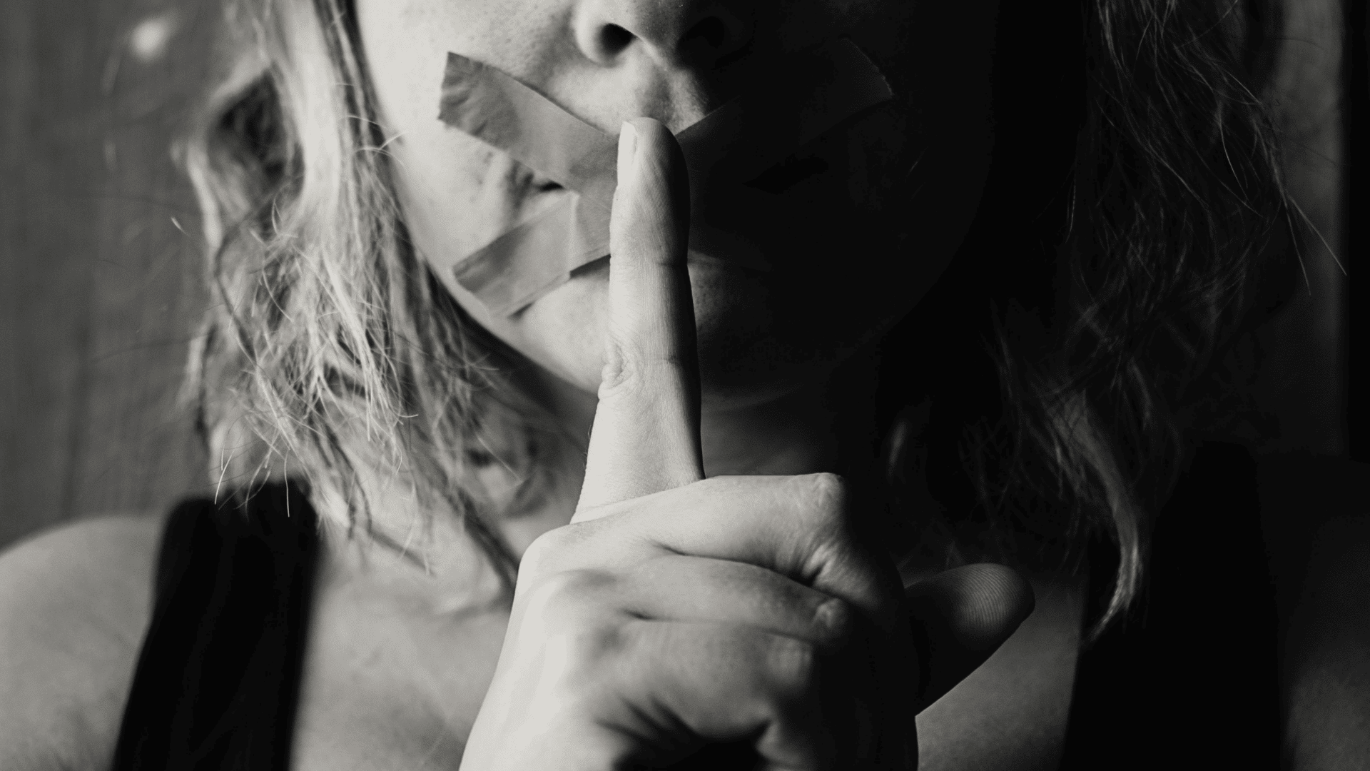 Woman with tape over her mouth making a silence gesture