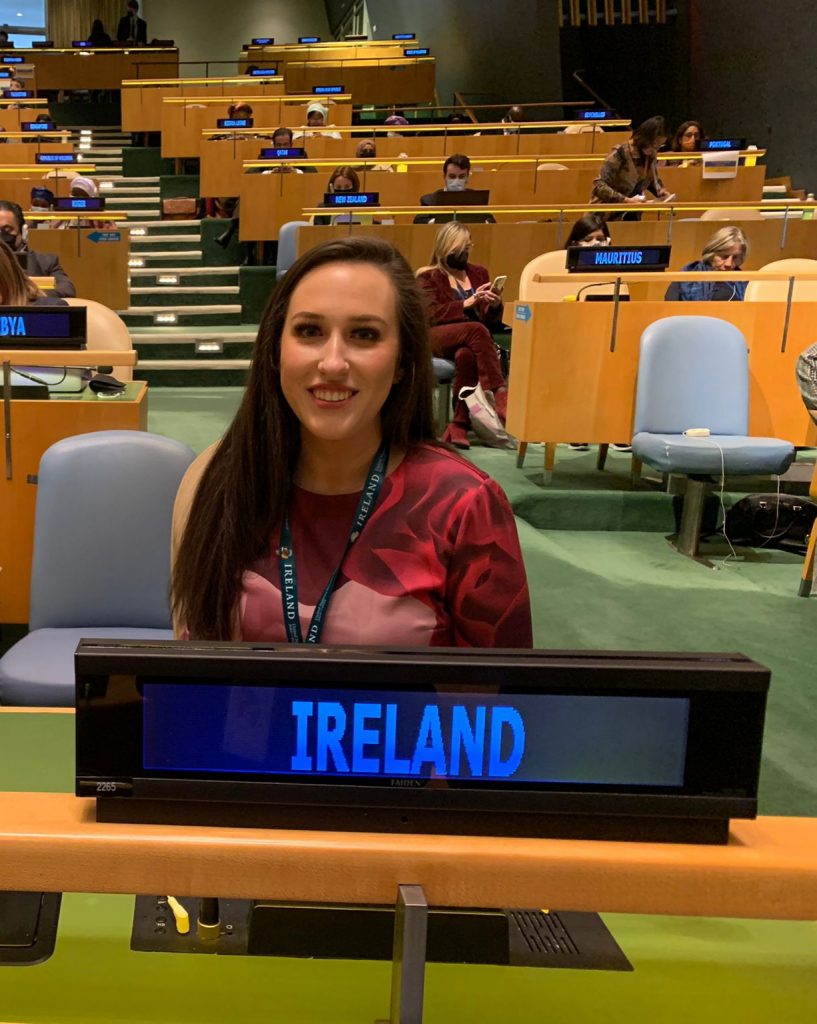 Diandra sits in the centre of the photo with a sign on a table in front of her which reads "Ireland". Behind is a large conference room with rows of tables and desks with other representatives at the 66th Session of the Commission on the Status of Women..