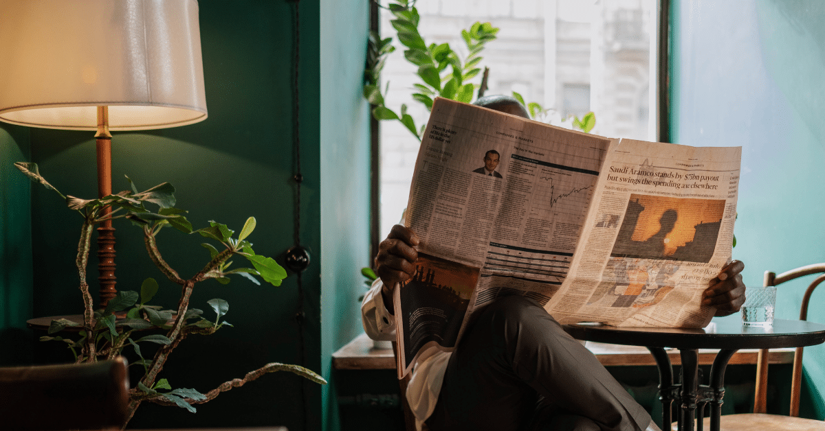 A photo of someone reading a newspaper inside a cosy cafe. Their face is obscured by the newspaper and their legs are crossed.