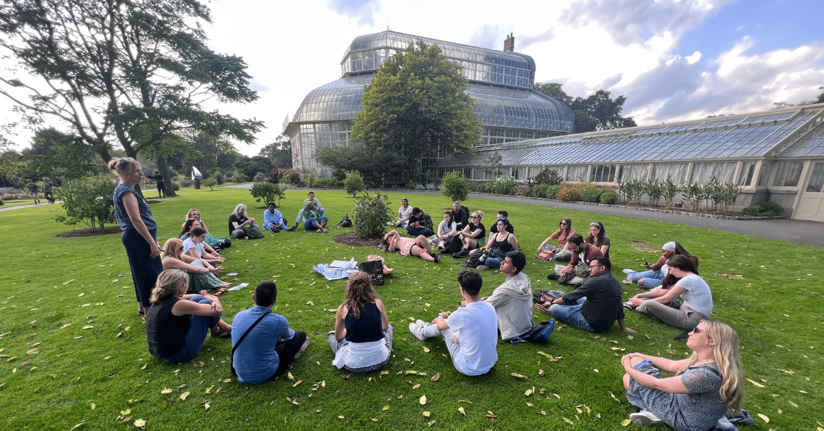 A photo of a group of people sitting in a circle on the grass in Dublin's botanical gardens. There is a large greenhouse behind them.