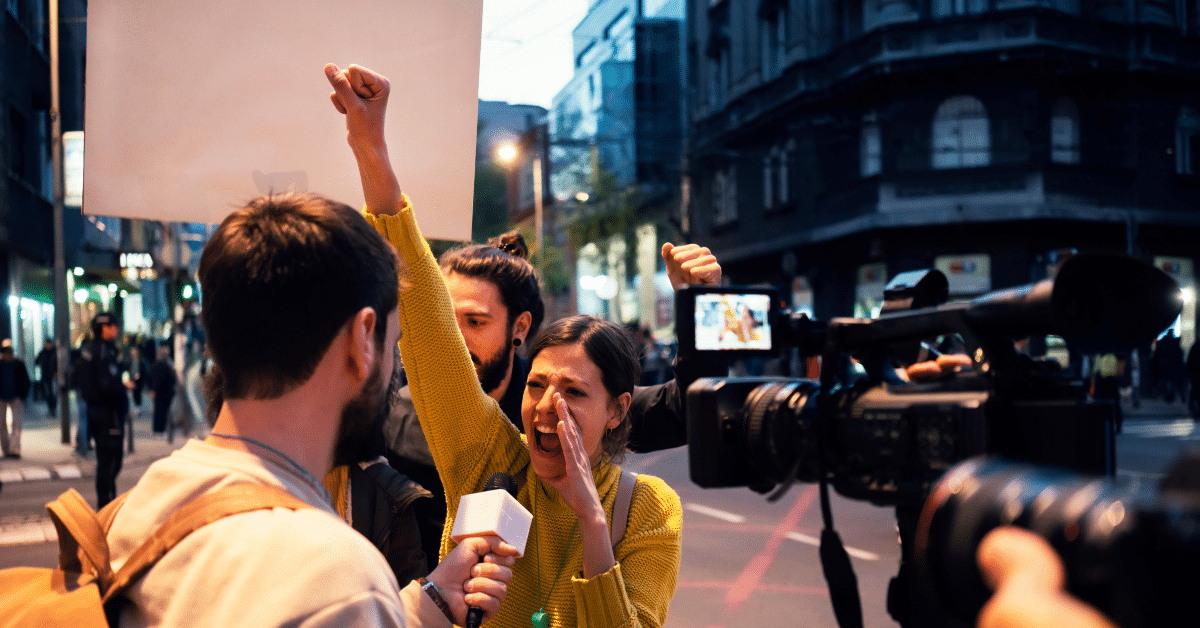 A photo of a young female protester shouting with her hand raised in a fist. She is being interviewed by a man with a microphone, and a news camera is facing her. There are other protesters with signs behind her.