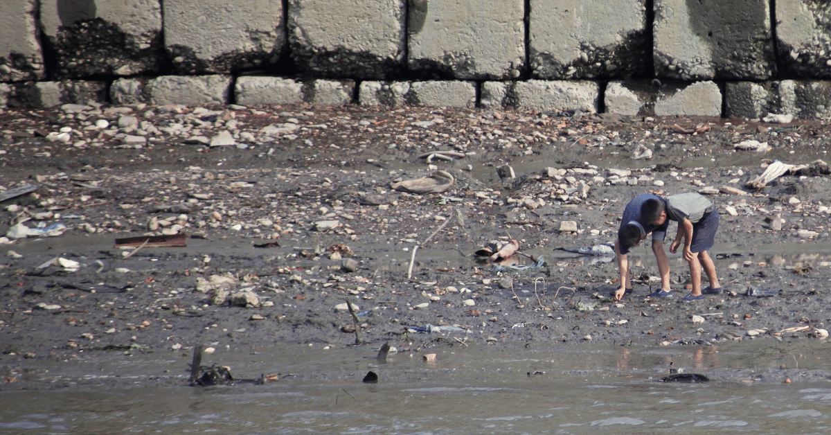 A photo of two small Korean children bent over, searching for food in the sand on a garbage-covered beach. The beach is very grey, and there's a big grey wall behind them.