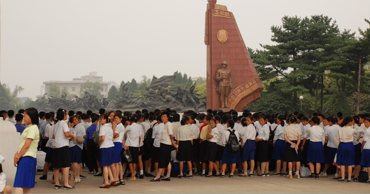 A group of between 50 and 100 Korean students stand in matching uniforms, looking at the Victorious Fatherland Liberation War Monument in Pyongyang, a series of large grey statues and a North Korean flag.