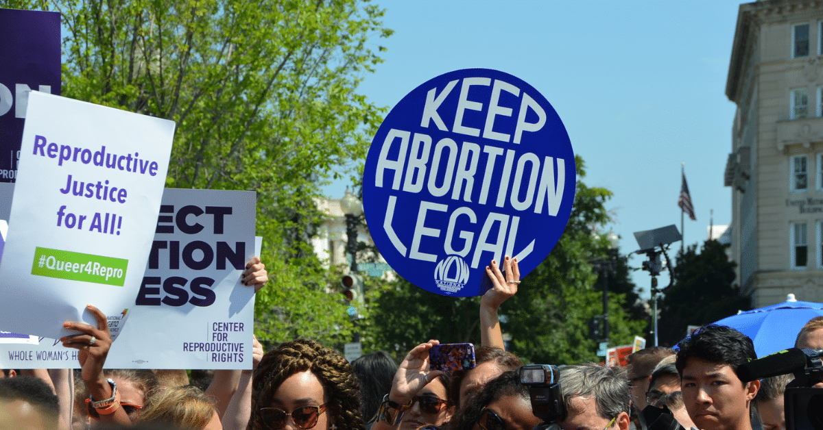 A photo of people holding various pro-abortion signs at a protest in Texas. The sky is blue and there are trees in the background. The most prominent sign in the centre reads: Keep abortion legal. Another to the left says: Reproductive Justice for All! #Queers4Repro